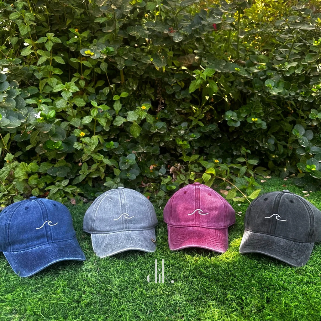 Four baseball caps in blue, gray, pink, and black on grass with a green bush background