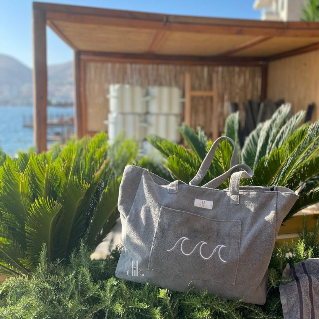 Gray tote bag with wave design on green plants by a wooden structure and body of water.
